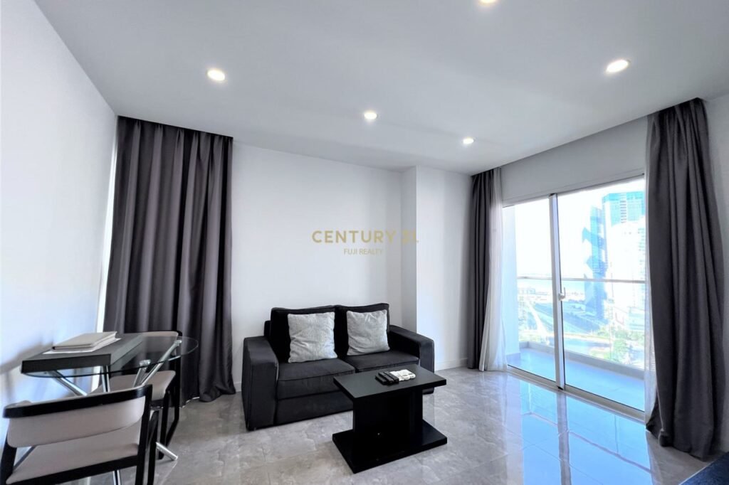 Modern 1 bedroom apartment living room with a black sofa, two white pillows, black coffee table, glass dining table with chairs, and large windows with gray curtains overlooking a city view. Bright white walls and tiled floor complete the open space.