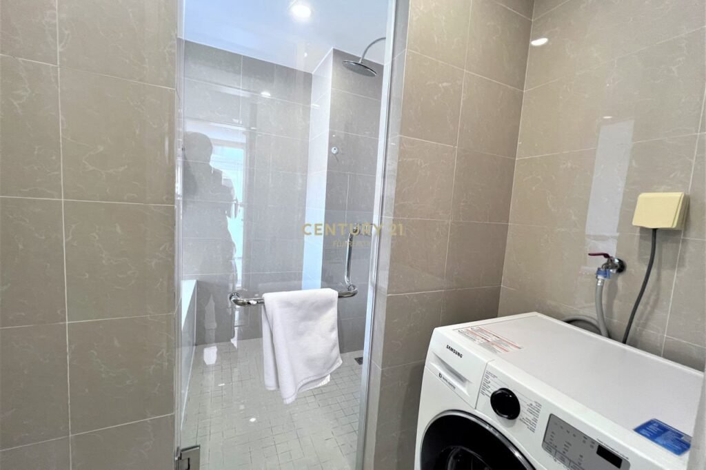 A modern Apartment 1 Bedroom bathroom with beige tiled walls, a glass shower stall with a towel on the handle, and a white washing machine beside a water connection. A faint reflection of a person is visible in the shower glass.