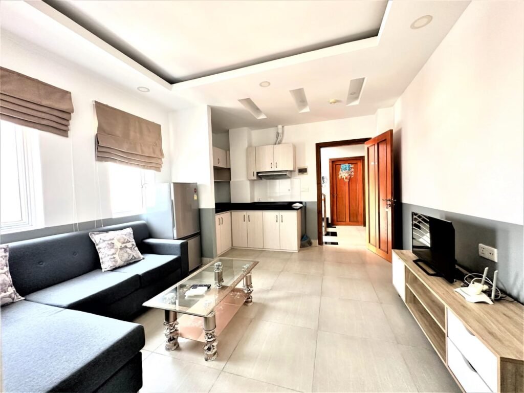 A bright living room in Condo L Olympic features a gray sectional sofa, glass coffee table, flat-screen TV on a stand, and an open kitchenette. Sunlight streams through windows with beige Roman shades.