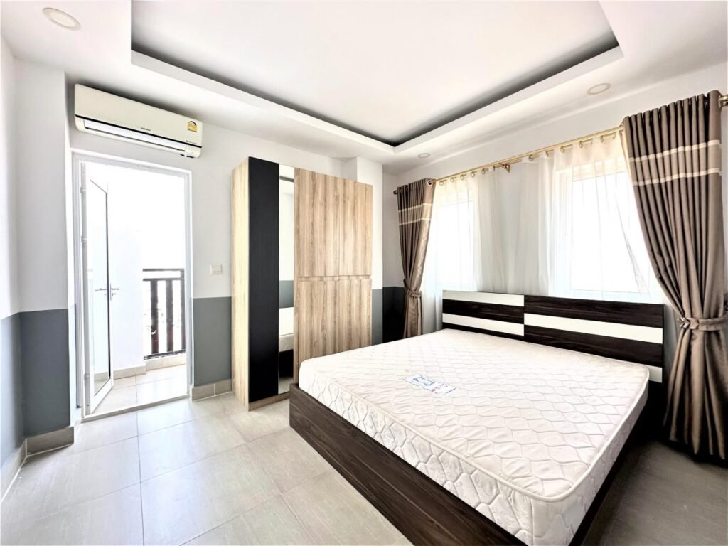 A bright, modern bedroom at Condo L Olympic features a large bed, a wooden wardrobe with mirrors, light tiled floor, brown curtains, and an air conditioner above a door that opens to a sunny balcony. Sunlight streams through the windows.