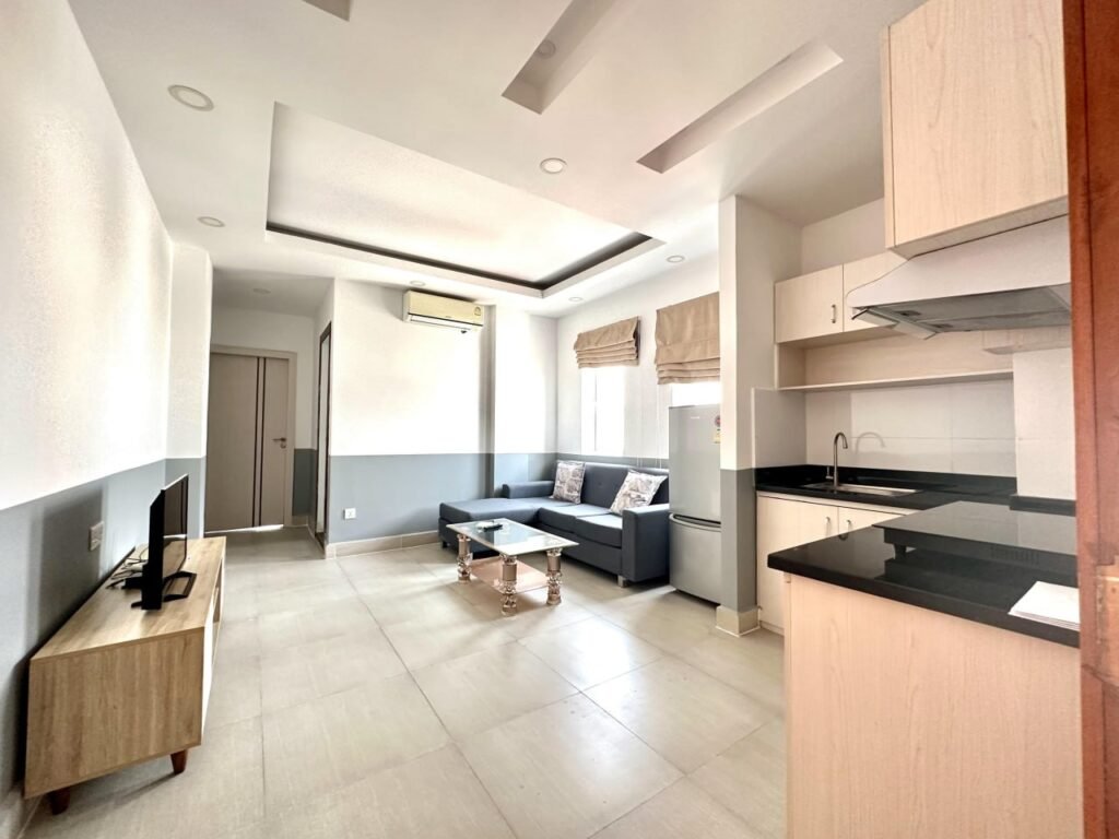 Bright, modern Condo L Olympic living room and kitchenette with light tile floors, L-shaped gray sofa, glass coffee table, flat-screen TV on a wooden stand, and white cabinetry under recessed ceiling lights.