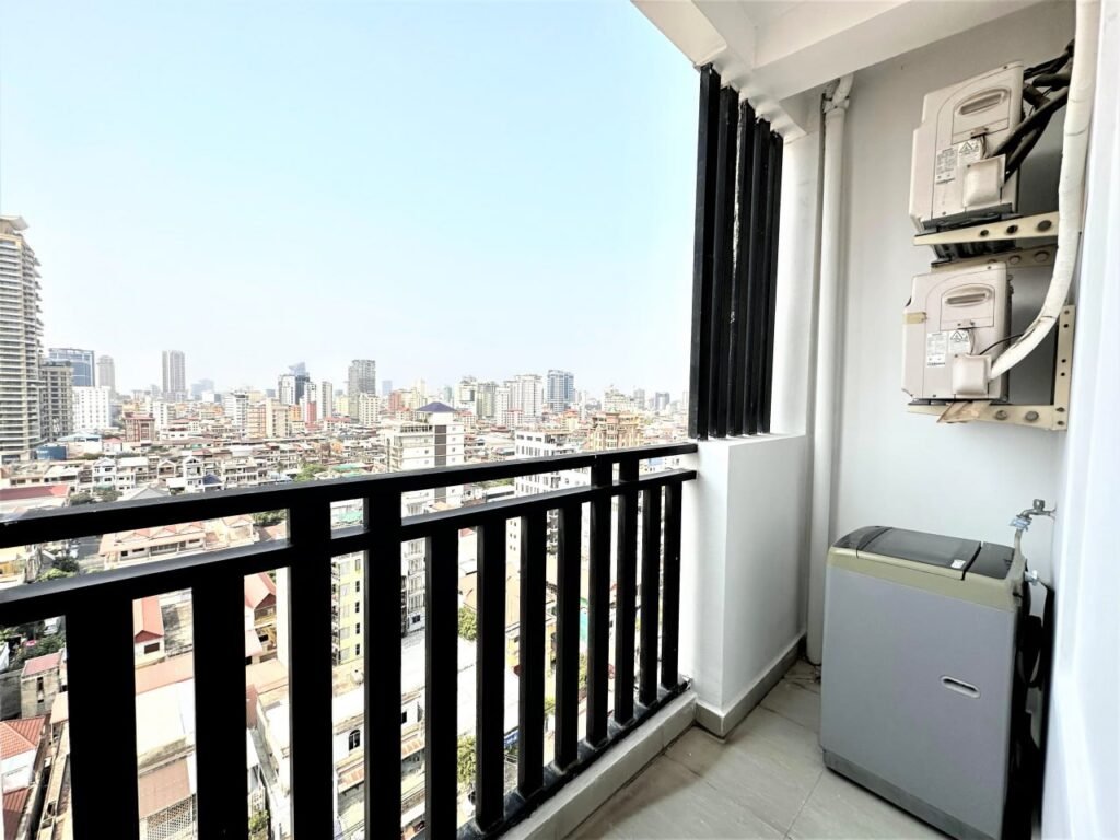 A high-rise Condo L Olympic balcony with black railings overlooks a city skyline. On the right, there’s a washing machine and utility meters mounted on the wall. The sky is clear and bright.