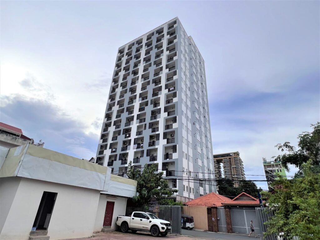 A tall, modern white Condo L Olympic apartment building stands against a partly cloudy sky, with a few smaller buildings, a white truck, and trees in the foreground.