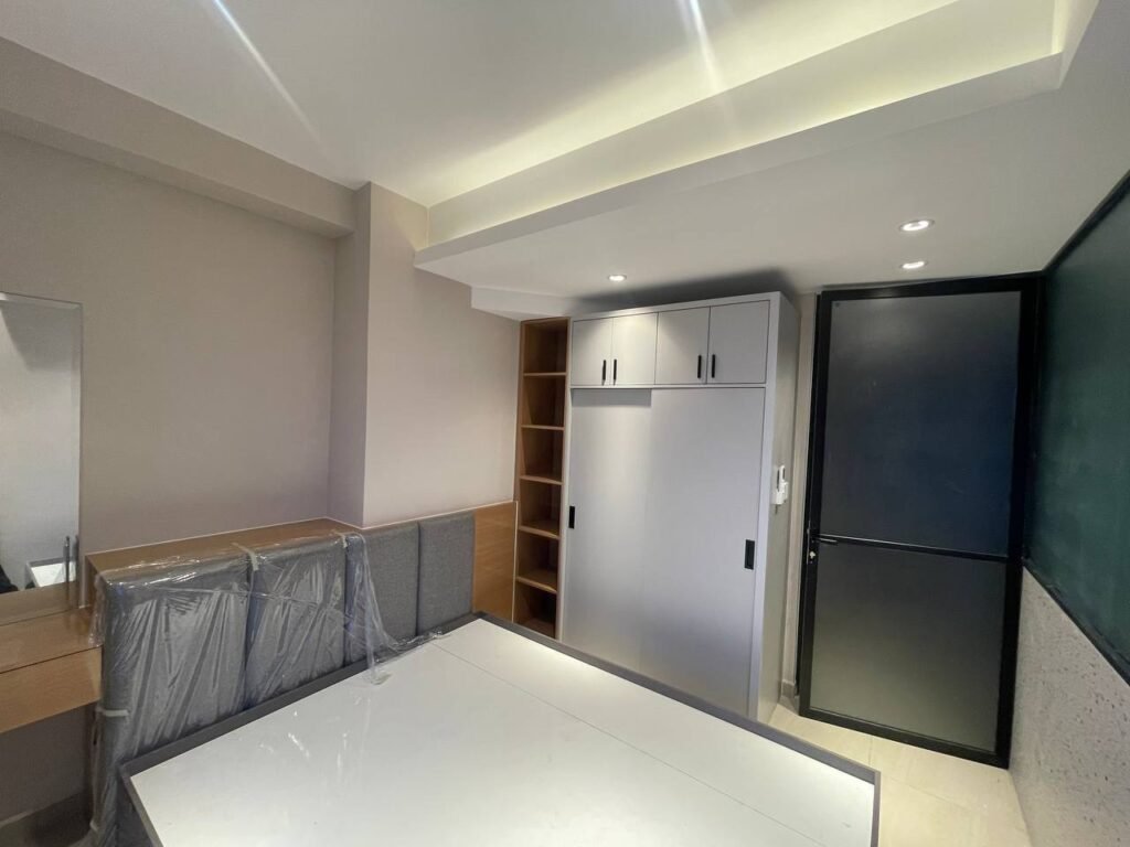 A modern, minimal L-Residence 2 Bedroom featuring a bed frame wrapped in plastic, a built-in wardrobe, open shelves, a black door, mirror, and recessed ceiling lights. The neutral decor includes light-colored walls and floors.