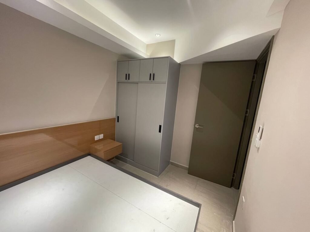 A small, minimal L-Residence 2 Bedroom with light-colored walls, a wooden headboard, simple bed frame, built-in nightstand, closed gray wardrobe, and closed gray door. The room appears clean and unfurnished.