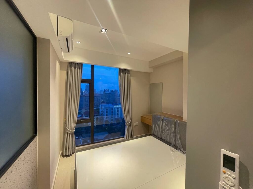 A modern, unfurnished L-Residence 2 Bedroom with large windows, beige walls, and gray curtains. A cityscape is visible outside. Features include a covered desk, wall-mounted air conditioner, and a remote control near the entrance.