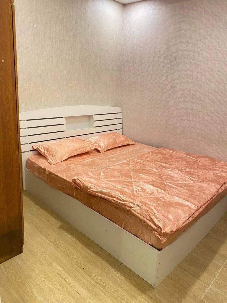 A neatly made bed with a white frame and peach-colored bedding, including two pillows and a comforter, sits in the corner of an L-Residence 2 Bedroom with light brown walls and a wooden floor.