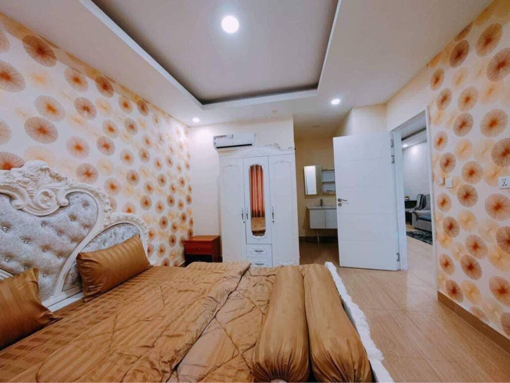 A cozy L-Residence 2 Bedroom features orange-patterned wallpaper, a tufted white headboard, matching brown bedding, a red bedside table, a white wardrobe, and an open door leading to a well-lit hallway and another room.