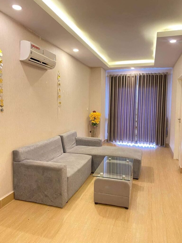 A modern L-Residence 2 Bedroom living room with a gray sectional sofa, glass coffee table, floor vase with yellow flowers, wooden flooring, beige walls, air conditioner, and floor-to-ceiling curtains covering a window or glass door.
