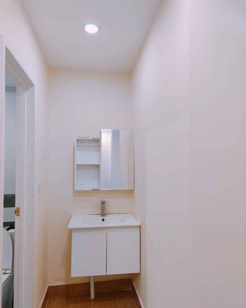 A small, narrow bathroom in the L-Residence 2 Bedroom features cream-colored walls, a white vanity with storage, a square sink, and a mirrored cabinet. The wood-toned floor adds warmth, with a doorway to the left for easy access.