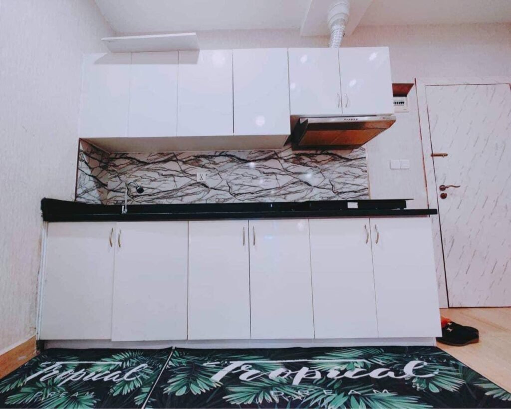 A modern L-Residence 2 Bedroom kitchen features white cabinets, black countertop, a stainless steel range hood, and a wavy black-and-white backsplash. Two mats with tropical leaf designs add a fresh touch to the floor.