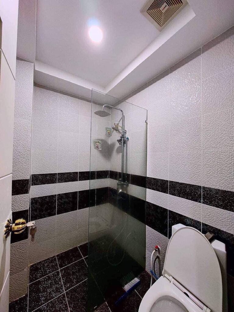 A modern L-Residence 2 Bedroom bathroom with a glass-enclosed shower, overhead showerhead, tiled walls featuring black horizontal stripes, and a white toilet with a bidet spray in the foreground.