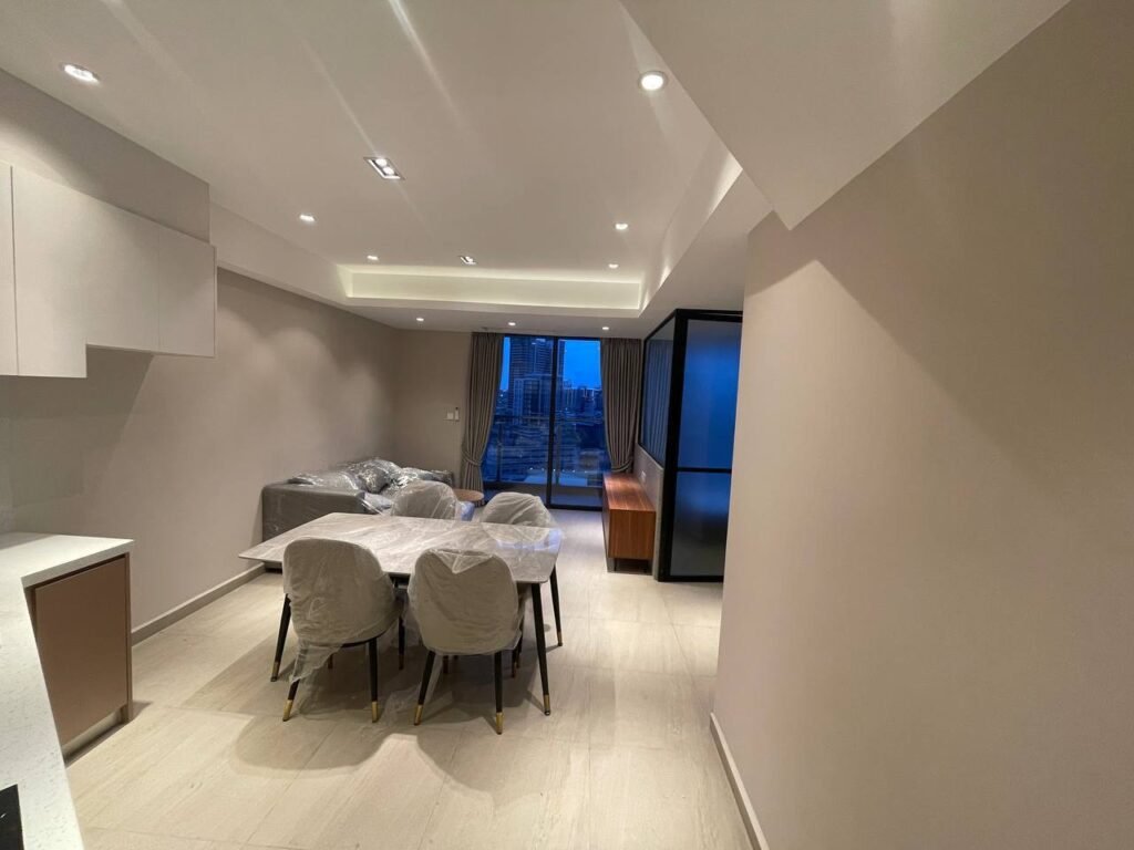 Modern L-Residence 2 Bedroom apartment interior with a dining table and six chairs, a living area featuring a sofa covered in plastic, large windows showcasing city views, and soft ceiling lighting in a neutral color palette.