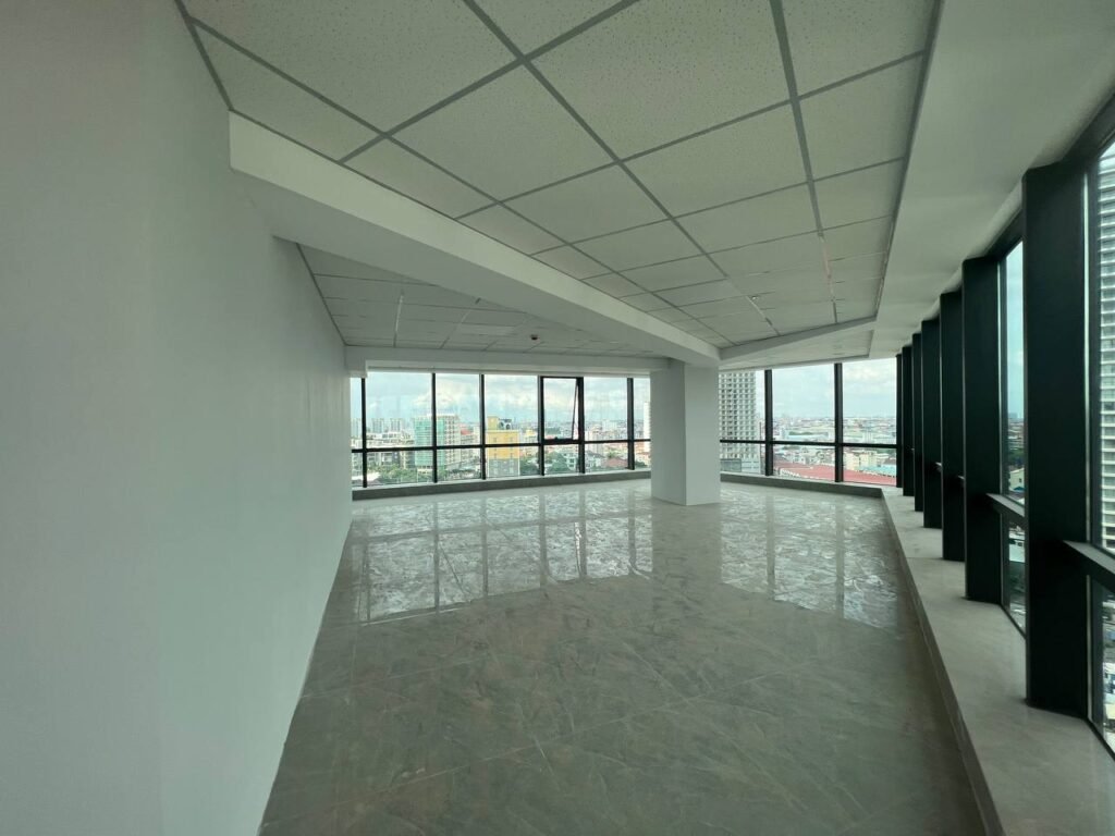 Spacious office space for sale featuring tiled floors, white walls, and a grid-pattern ceiling. Large floor-to-ceiling windows provide stunning cityscape views with buildings and a partly cloudy sky.