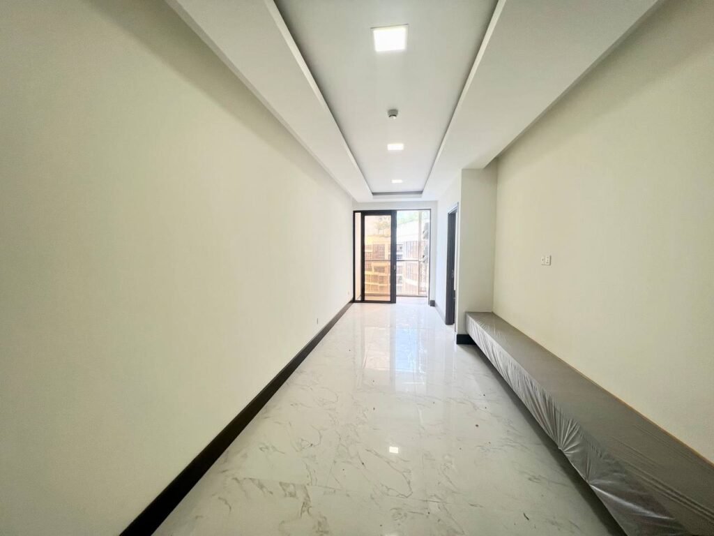 A bright, empty room at Orkide The Royal boasts white walls and a shiny marble floor, with recessed ceiling lights, a long narrow window, and a glass door to the balcony. A built-in bench lines one wall for added style.
