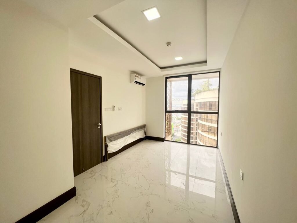 A bright, empty room at Orkide The Royal features white marble floors, white walls, a dark wooden door, ceiling light, air conditioner, and large glass doors opening to a balcony with views of nearby buildings.