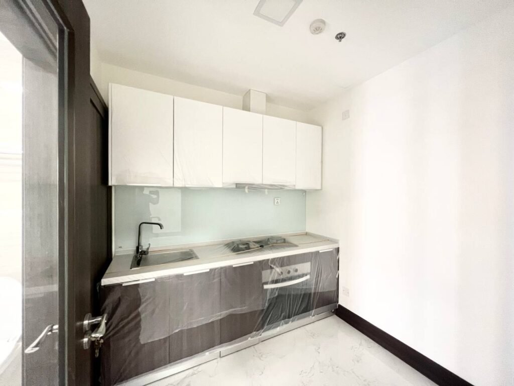 A small modern kitchen at Orkide The Royal features white upper cabinets, dark lower cabinets, a built-in cooktop and sink, plus a white marble floor. The cabinetry is partially covered in protective plastic. Bright space with crisp white walls.
