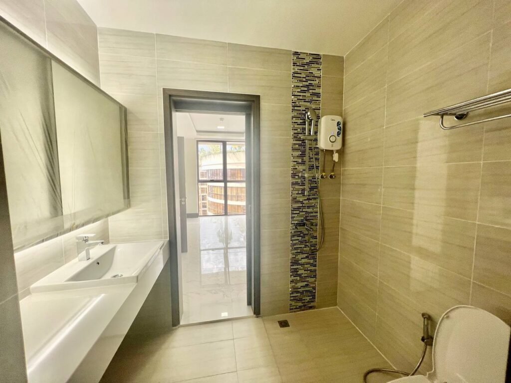 Modern bathroom at Orkide The Royal with beige tiles, a long mirror above a white sink, a walk-in shower with water heater and blue accent tiles, towel rack, and toilet. Natural light brightens the space from the glass door.