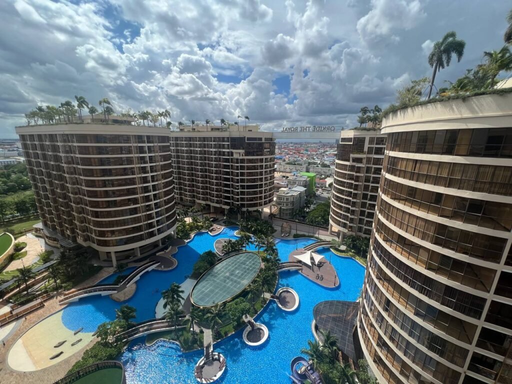 A high-angle view of several curved, modern high-rise buildings at Orkide The Royal surrounds a large, irregularly shaped swimming pool with palm trees and walkways. The cityscape and cloudy sky are visible in the background.