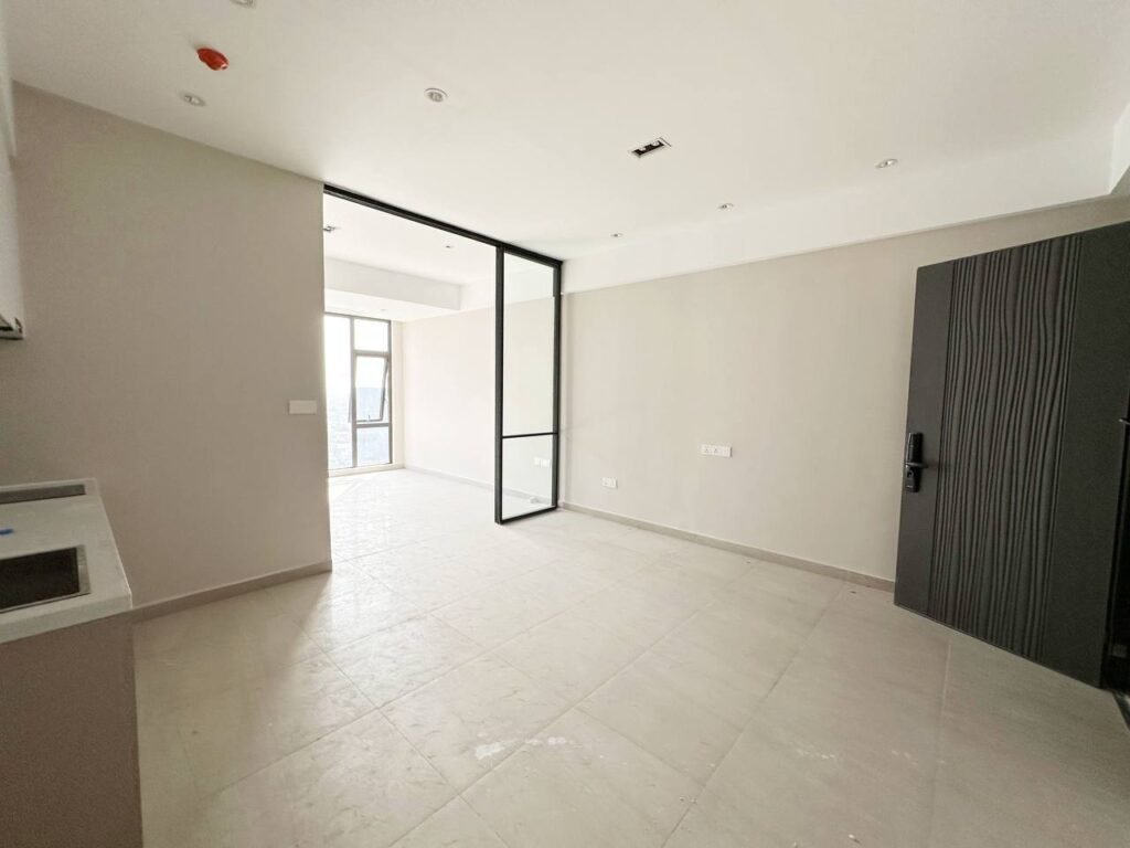 A bright, empty modern 1 bedroom apartment with light-colored tiled floors, white walls, a dark door, and a large glass partition leading to a small balcony with a glass door. Ceiling lights and a kitchen sink are visible.