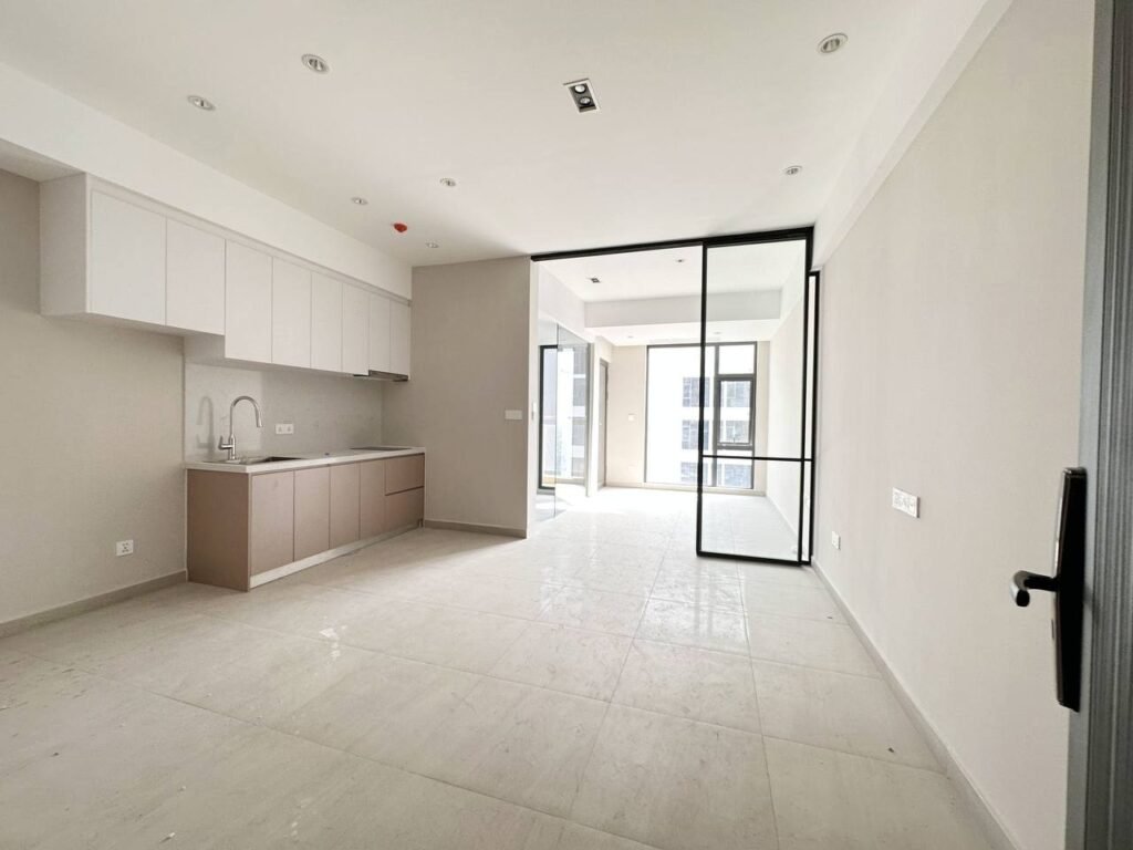 A modern, empty 1 bedroom apartment featuring light-colored walls and floors, a small kitchen area on the left, and a glass door leading to a bright room with large windows.