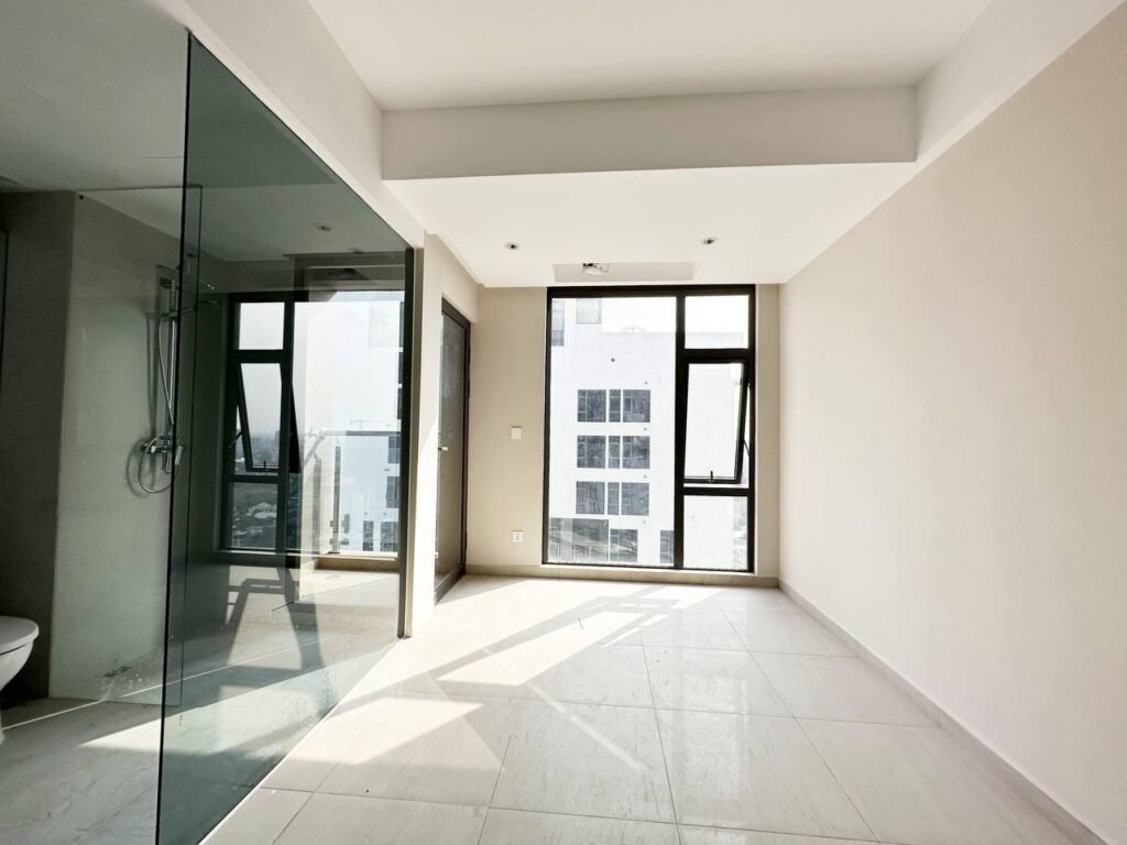 A bright, empty 1 bedroom apartment with large floor-to-ceiling windows, light-colored tiled floors, and a glass-enclosed shower area on the left. Sunlight streams in, casting soft shadows across the spacious floor.