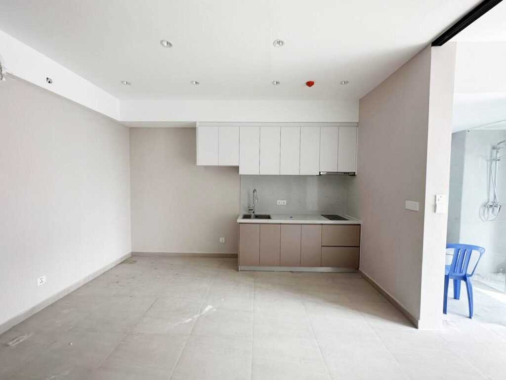 Minimalist kitchen in a 1 bedroom apartment with white upper cabinets and beige lowers, gray countertop, built-in sink, tiled flooring, off-white walls, and a blue plastic chair near a glass partition.