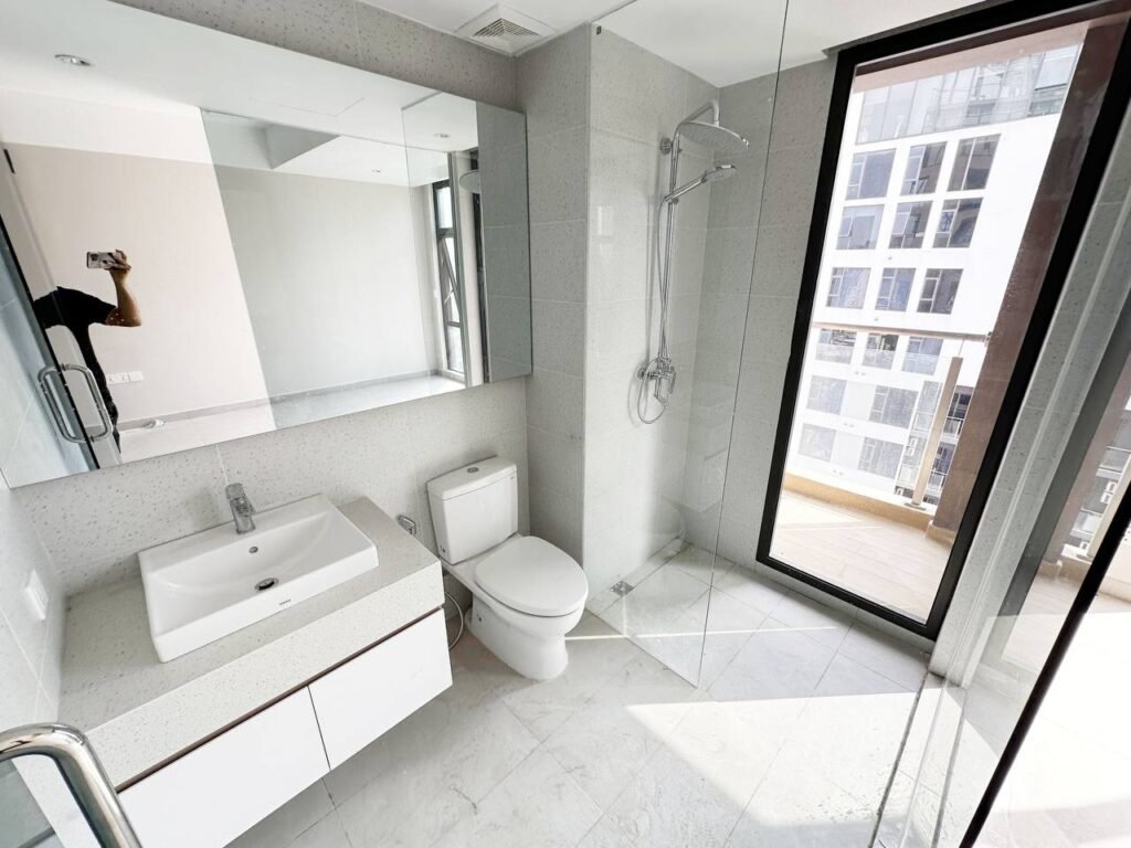A modern bathroom in a 1 bedroom apartment with a white sink, toilet, and glass-enclosed shower. A large mirror sits above the sink, while a floor-to-ceiling window reveals city buildings. The space is bright and minimalistic.