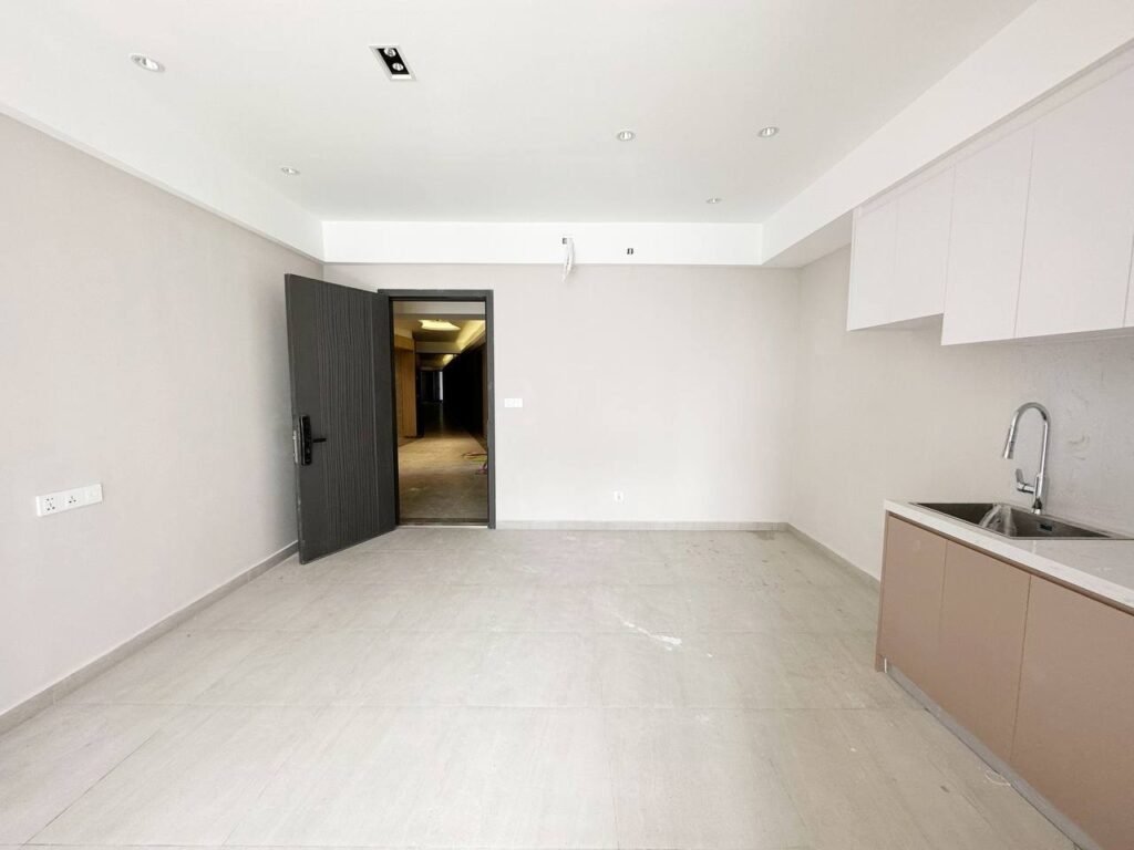 A minimal, empty 1 bedroom apartment with light-colored walls and tile flooring, featuring an open dark door, a small kitchenette with a sink and cabinets, and recessed ceiling lights.