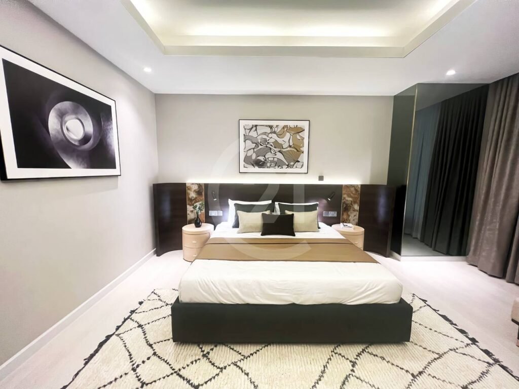 A modern bedroom in a 3 bedroom apartment features a large bed with neutral bedding, two round bedside tables, abstract art on the walls, a patterned rug, and soft, warm lighting beneath floor-to-ceiling curtains.