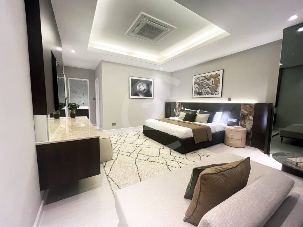 A modern, spacious bedroom in a 3 bedroom apartment features a large bed, abstract wall art, neutral tones, a patterned rug, cozy sofa, and built-in storage. Recessed ceiling lighting adds to the warm, inviting atmosphere.