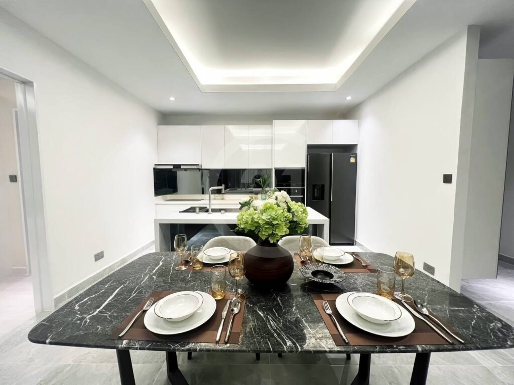 Modern kitchen and dining area in a 3 bedroom apartment, featuring a black marble table set for four with gold-rimmed glasses, white plates, and green hydrangeas; sleek white cabinets and appliances complete the elegant space.