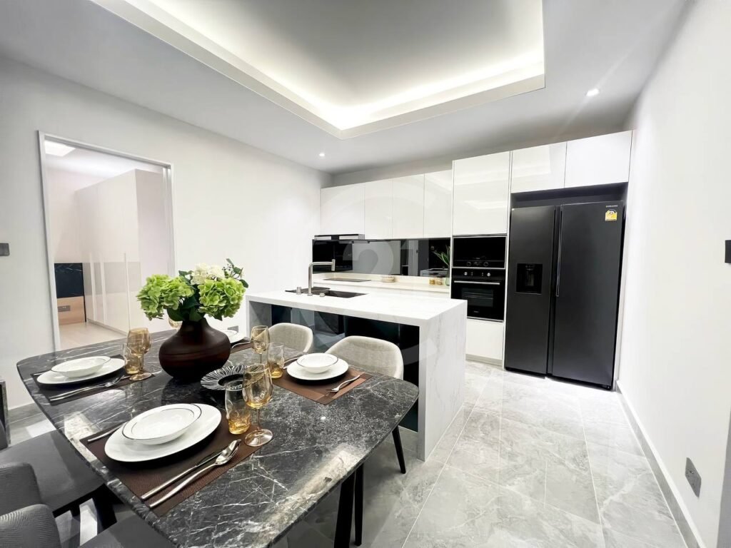 Modern kitchen and dining area in a 3 bedroom apartment, featuring a marble dining table set for four, an island with bar stools, white cabinets, built-in oven, and a large black refrigerator. The space is bright with sleek, minimalist decor.