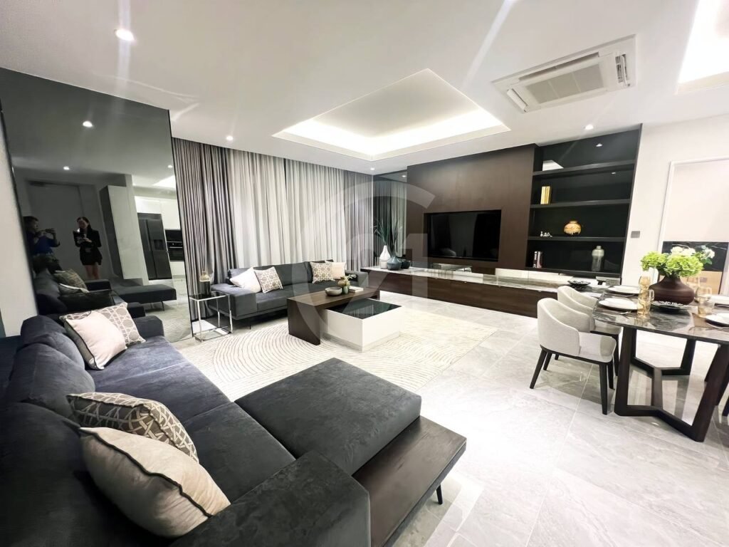 Modern 3 bedroom apartment living room with dark sectional sofa, glass coffee table, TV on a wood-paneled wall, shelves with decor, round dining table and chairs, plus large windows with gray curtains. Two people can be seen reflected in a wall mirror.