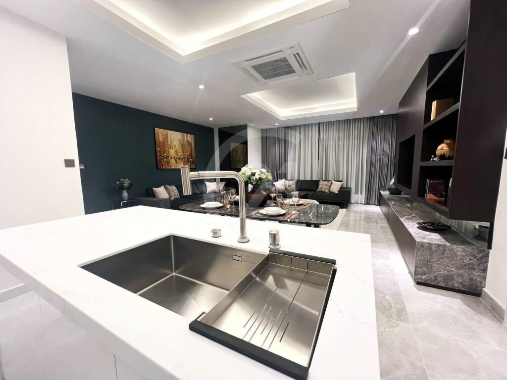 Modern 3 bedroom apartment featuring a marble kitchen island and sink in the foreground, a dark sofa and coffee table, wall art, TV on a sleek unit, and floor-to-ceiling curtains covering large windows.