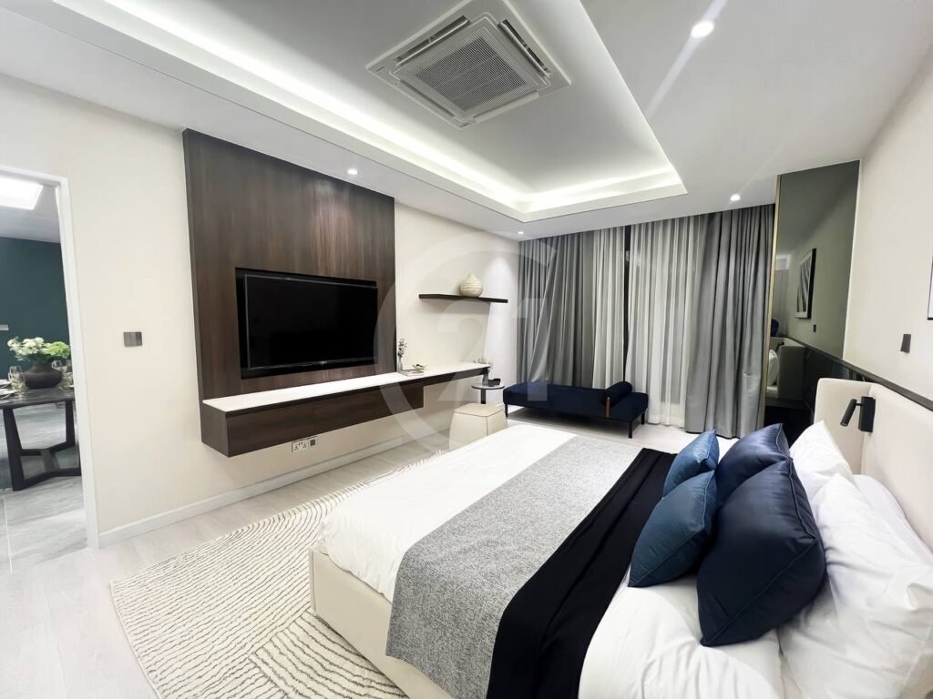 A modern 3 bedroom apartment features a spacious bedroom with a large bed, dark blue and black accent pillows, wall-mounted TV, floating shelf, stylish rug, tall curtains, small seating area, and an open doorway leading to the dining room.