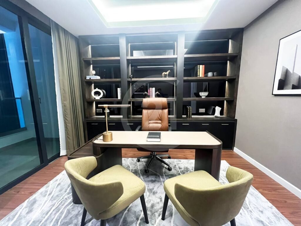 A modern office in a 3 bedroom apartment features a large bookshelf, brown desk chair behind a sleek desk, two light green chairs in front, a desk lamp, and rug. Floor-to-ceiling windows and framed art decorate the space.