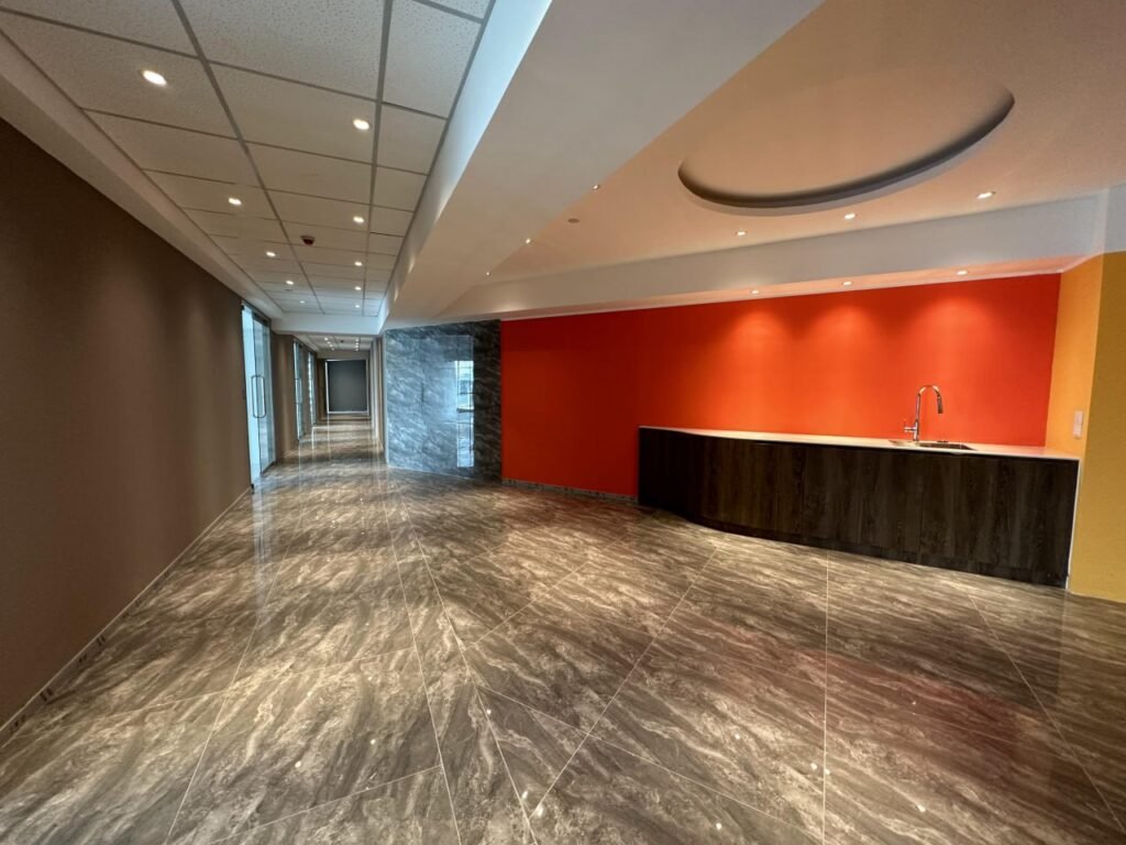Modern hallway with glossy marble floors, a dark wood kitchenette with a sink, and a bright orange accent wall. The corridor extends to glass doors, with recessed lighting in the ceiling.