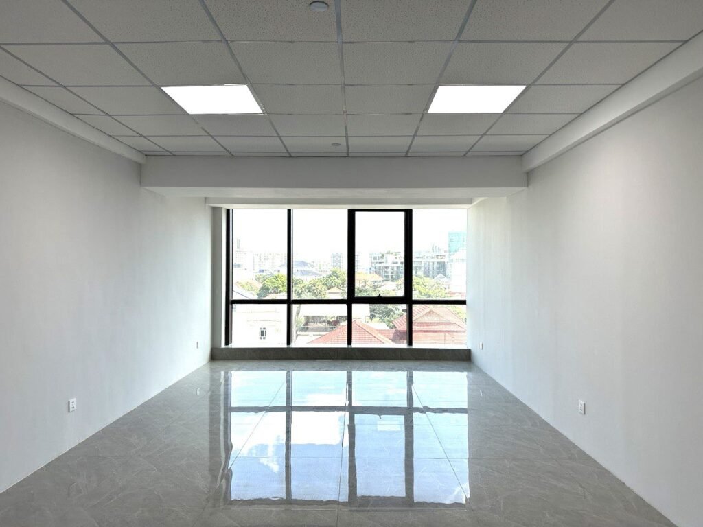 A bright, empty room with white walls, a tiled floor, a drop ceiling with square lights, and large windows letting in sunlight and offering a city view.