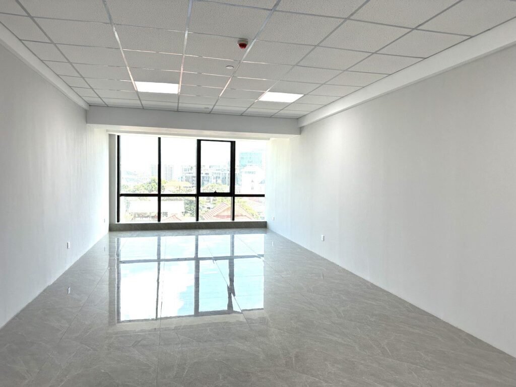 A bright, empty room with white walls, a tiled floor, and a large window letting in natural light. The window offers a view of buildings and trees outside. The ceiling has recessed lights and a fire alarm.