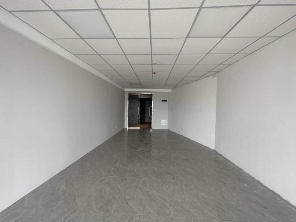 Empty, narrow room with white walls, tiled ceiling, and gray marble-like floor; a door and a small window are visible at the far end of the space.