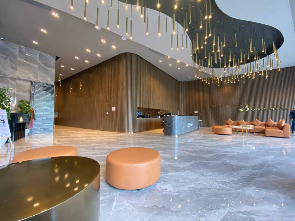 A modern hotel lobby with marble floors, brown leather seating, a curved wooden wall, a reception desk, and decorative hanging lights from the ceiling. Cozy lounge chairs and a gold table are also visible.