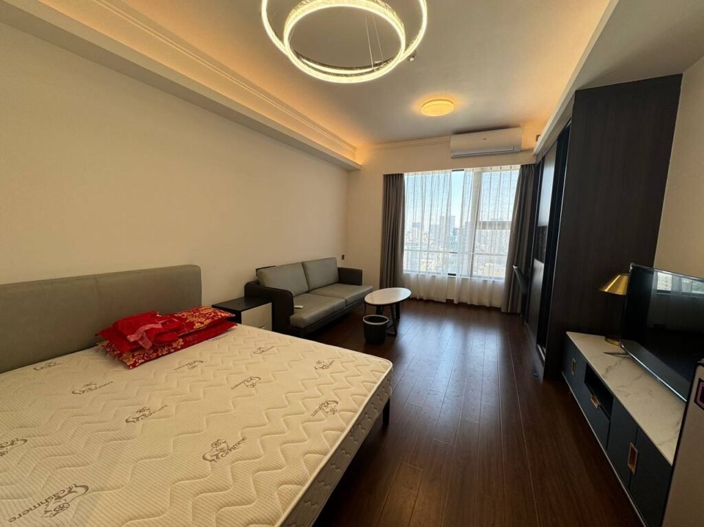 A modern bedroom with a large bed, gray headboard, and red pillows, a gray sofa, a round coffee table, a flat-screen TV on a dark cabinet, wood flooring, and large windows with sheer curtains letting in natural light.