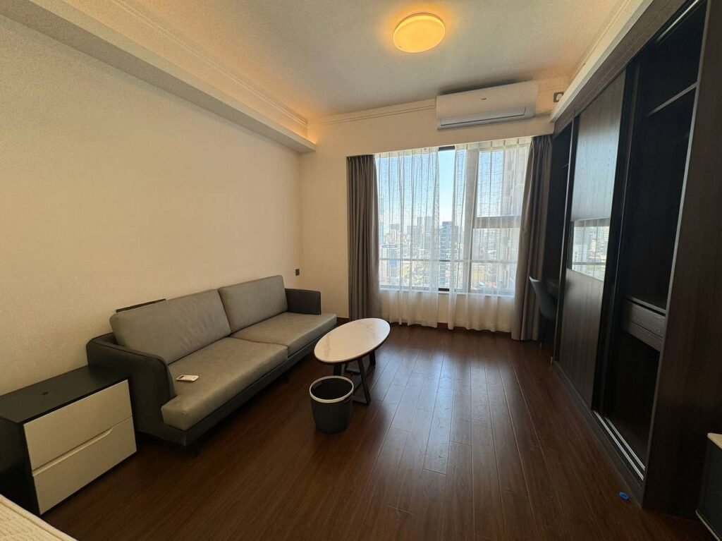 A modern, minimally furnished living room with a gray sofa, small white and black coffee tables, a nightstand, wooden floors, large window with sheer curtains, and city view outside.