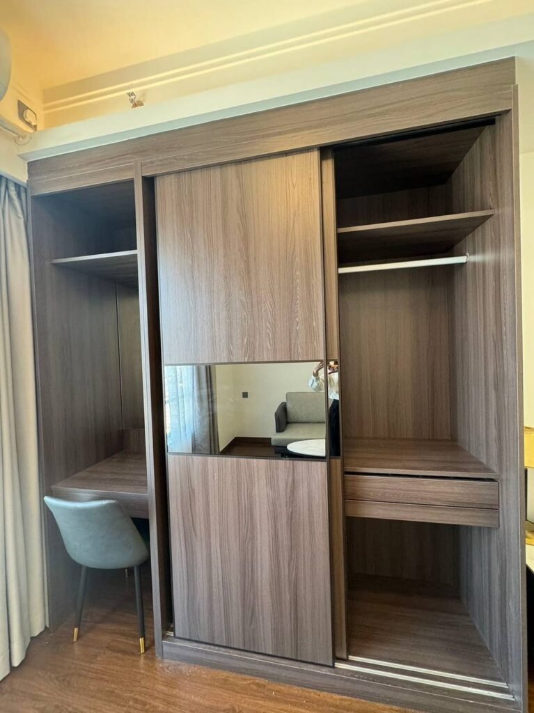 A modern wooden wardrobe with sliding doors, open shelves, and a built-in desk area with a gray padded chair. The wardrobe is placed against a wall in a well-lit room with wooden flooring.