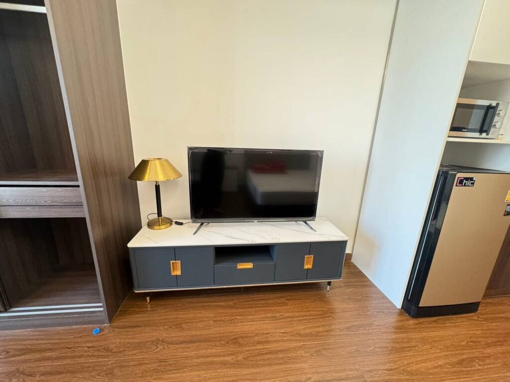 A flat-screen TV sits on a modern cabinet with gold handles, next to a gold table lamp. To the right is a small refrigerator, and to the left is a wooden wardrobe. The floor is wood and the walls are light-colored.