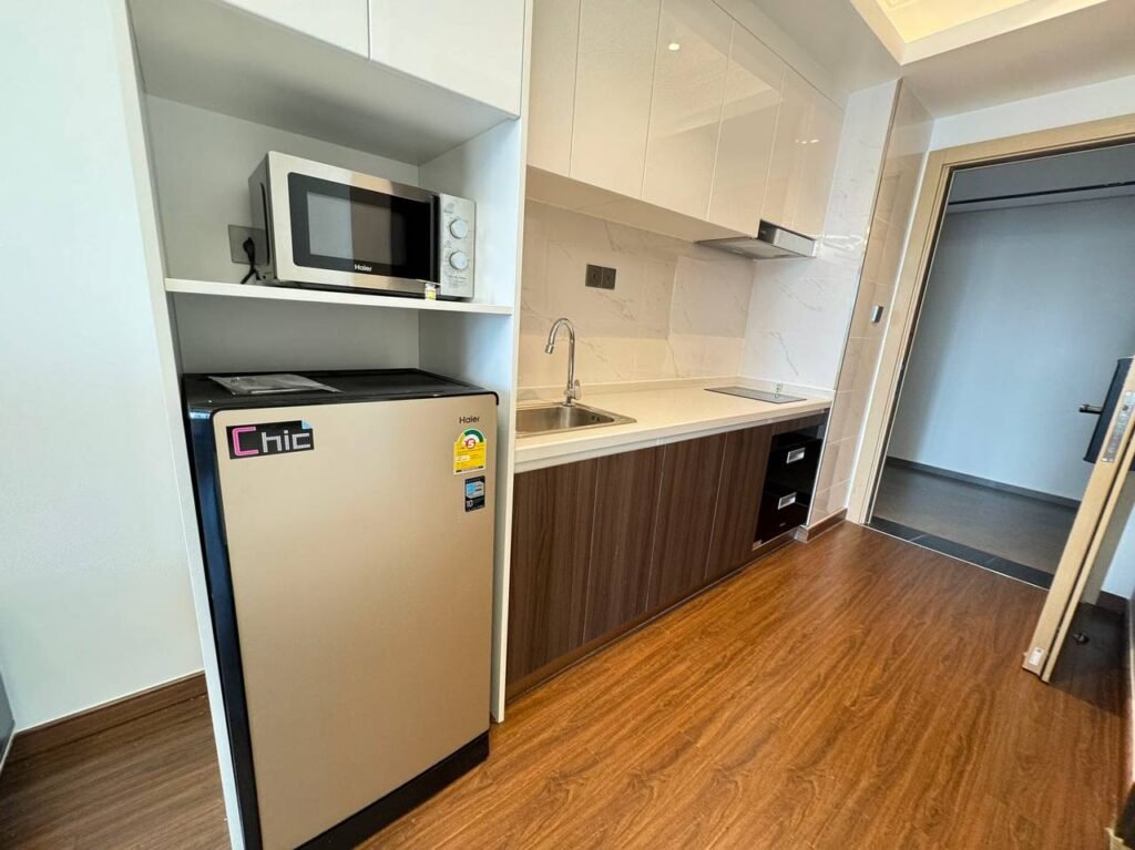 A compact modern kitchen with a small fridge, microwave, sink, cabinets, and built-in stovetop, featuring wood flooring and white countertops. An open door leads to another room.