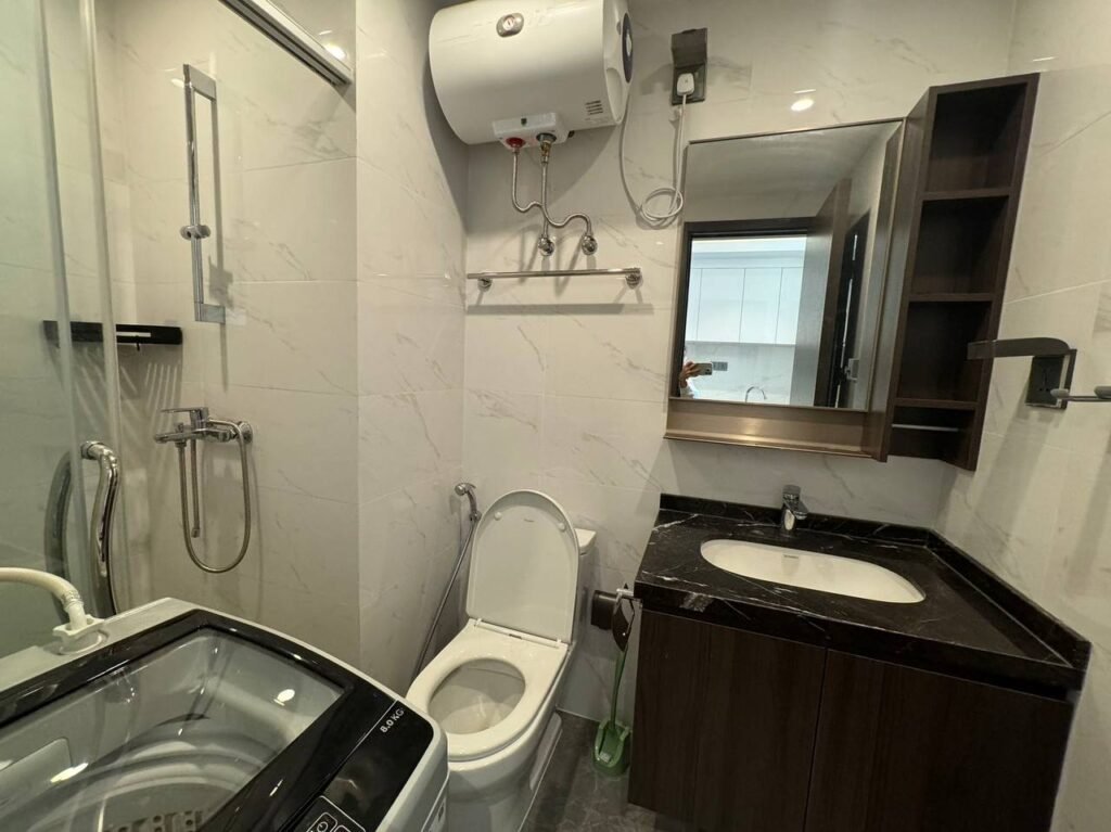 Modern bathroom with a washing machine, shower area with glass partition, toilet, wall-mounted water heater, sink with a black countertop, mirror cabinet, and shelves on white tiled walls.