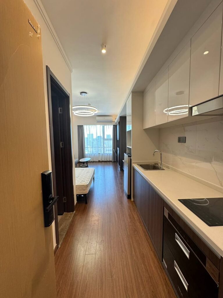 A modern studio apartment with wood flooring, a compact kitchen on the right, a bed near large windows in the background, and minimalistic decor. Light streams in through sheer curtains, creating a bright, cozy space.