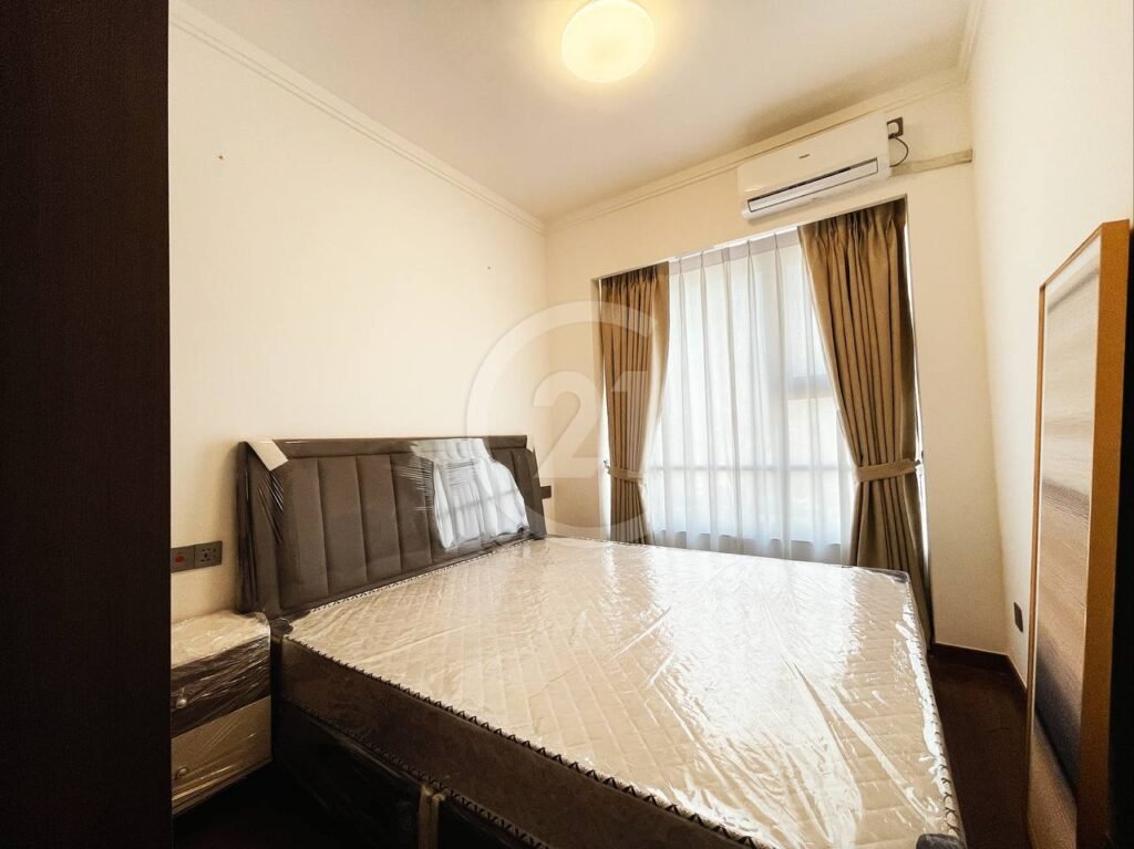 A small, minimally furnished bedroom with a double bed covered in plastic, a bedside table, brown curtains, a large window, and an air conditioner mounted above the window.